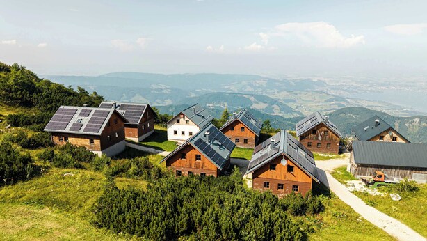Un ensemble de chalets en bois, dotés de panneaux solaires sur les toits, entourés de verdure et dominant un paysage montagneux.
