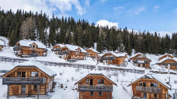 A cluster of wooden cabins with balconies, set on a snowy hillside with a forest backdrop.