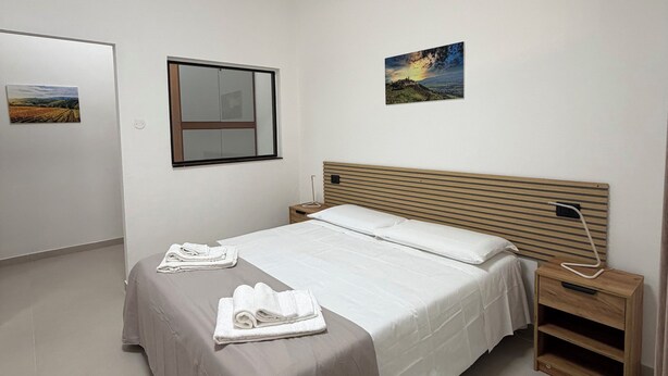 A hotel room with a bed, a bedside table, a nightstand, a wall-mounted lamp, and two framed pictures. - Guest House Colfiorito by Ospitaci-appartamento (Colfiorito)