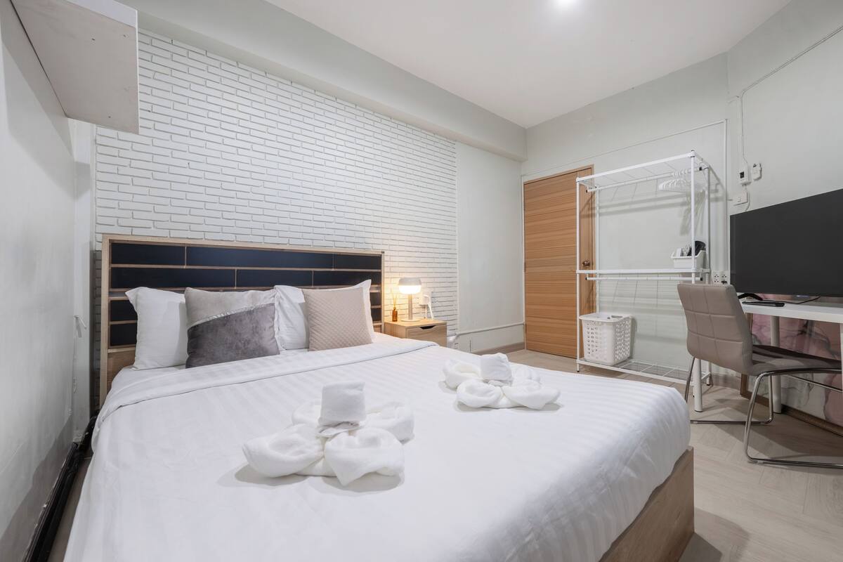 A hotel room with a white brick wall, a bed with white bedding and towels, a desk with a chair, a TV, and a wooden door.