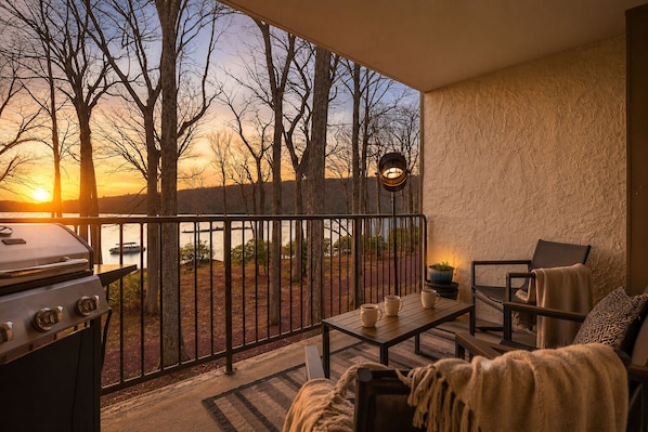 A balcony with a grill, a table with cups, a chair, and a view of trees and a sunset.