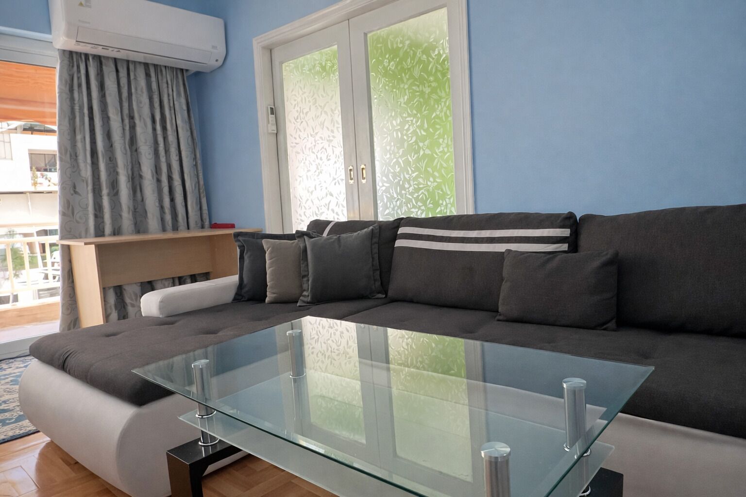 A modern living room with a glass coffee table, grey sofa, and a balcony with a wooden railing.
