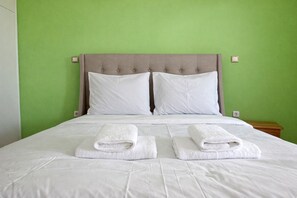 A bed with two pillows and two folded towels, a wooden nightstand, and a green wall. - Athens Getaway - Perfect Stay for 4 by ΜΡS (Athina)