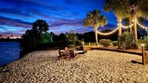 A beach setting with palm trees, string lights, and chairs. - Resort style 5-bedroom lake house with white sand beach, pool, and pool house. (Douglas)