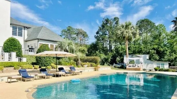 A swimming pool with lounge chairs, a covered area, and a building with a white facade and a sloped roof. - Resort style 5-bedroom lake house with white sand beach, pool, and pool house. (Douglas)