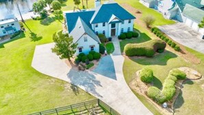 A large, two-story house with a prominent gable roof, surrounded by well-maintained lawns and a body of water. - Resort style 5-bedroom lake house with white sand beach, pool, and pool house. (Douglas)