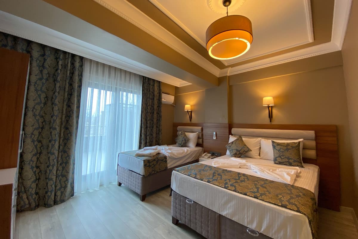 Una camera d'albergo con due letti, un'ampia finestra con tende leggere, un lampadario e lampade a parete.