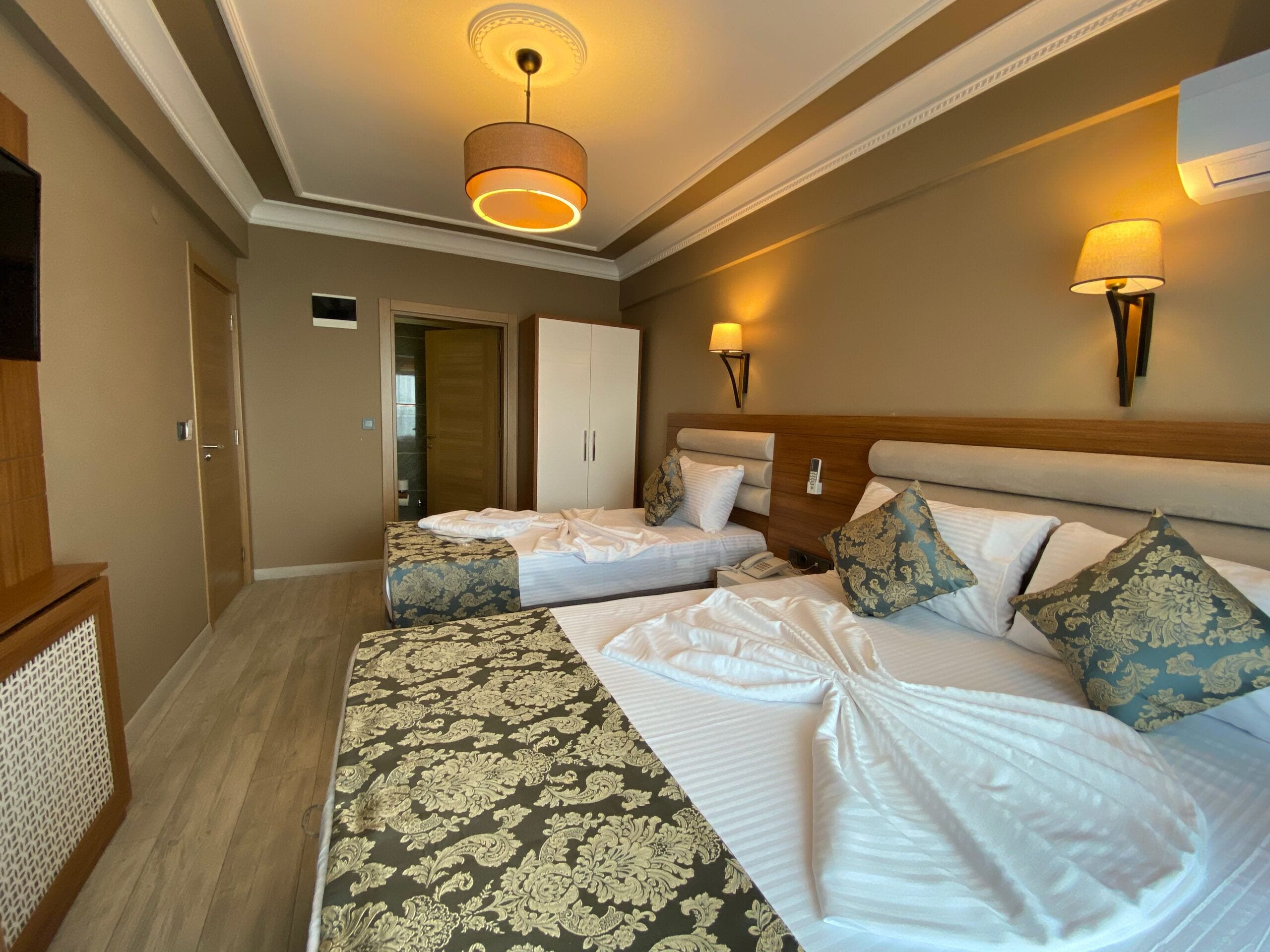 Habitación de hotel con dos camas, cabecero de madera, lámpara de pared y puerta que da a un baño.