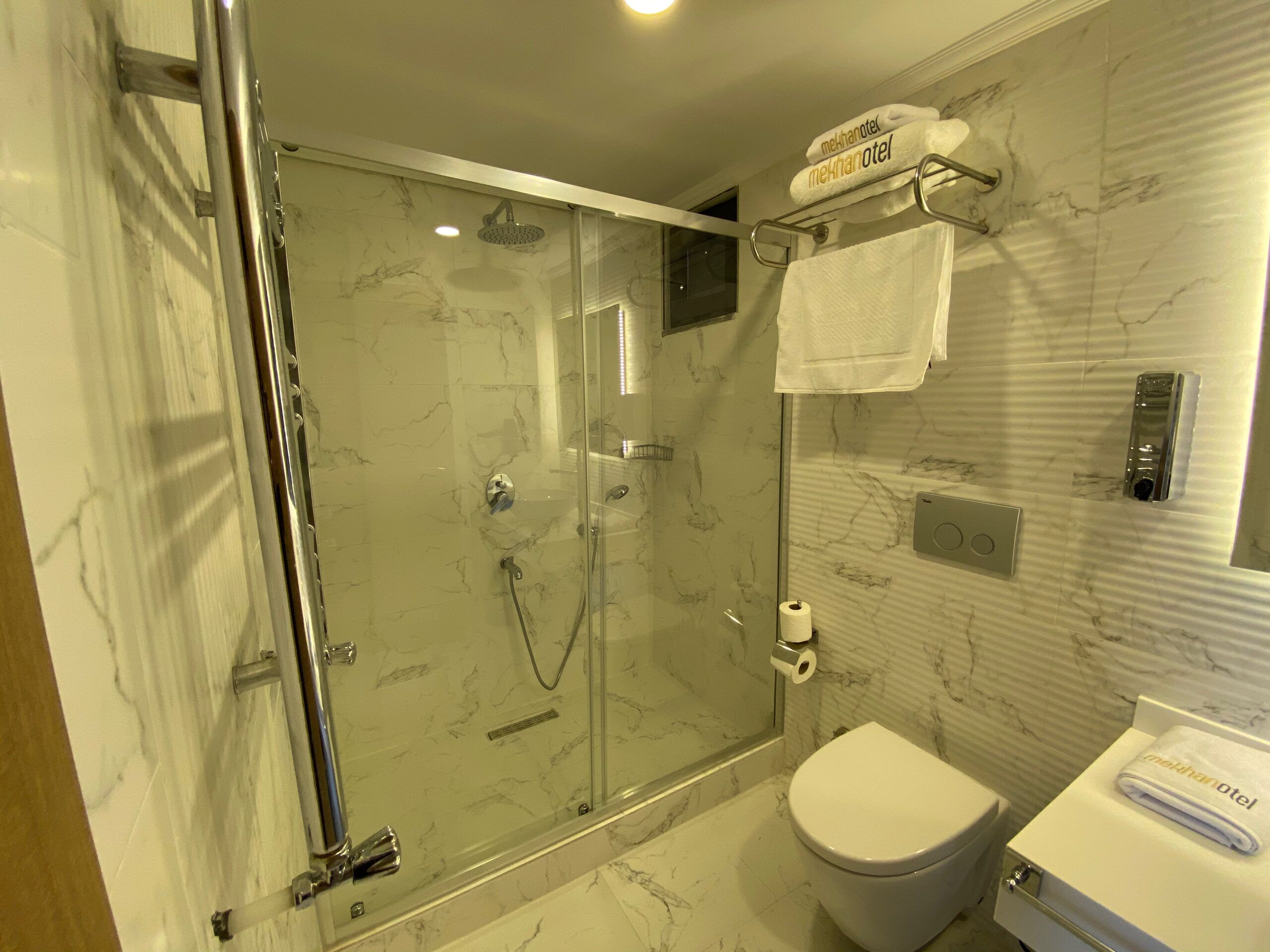 A modern bathroom with a glass-enclosed shower, a toilet, and a towel rack with a hotel name towel.