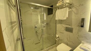Un baño moderno con ducha envuelta en vidrio, un inodoro y un perchero con una toalla que muestra el nombre del hotel.