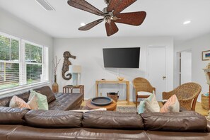 A living room with a brown leather sofa, a ceiling fan, a flat-screen TV, wicker chairs, and a fireplace. - Great home on the South end of St Simons (Saint Simons Island)