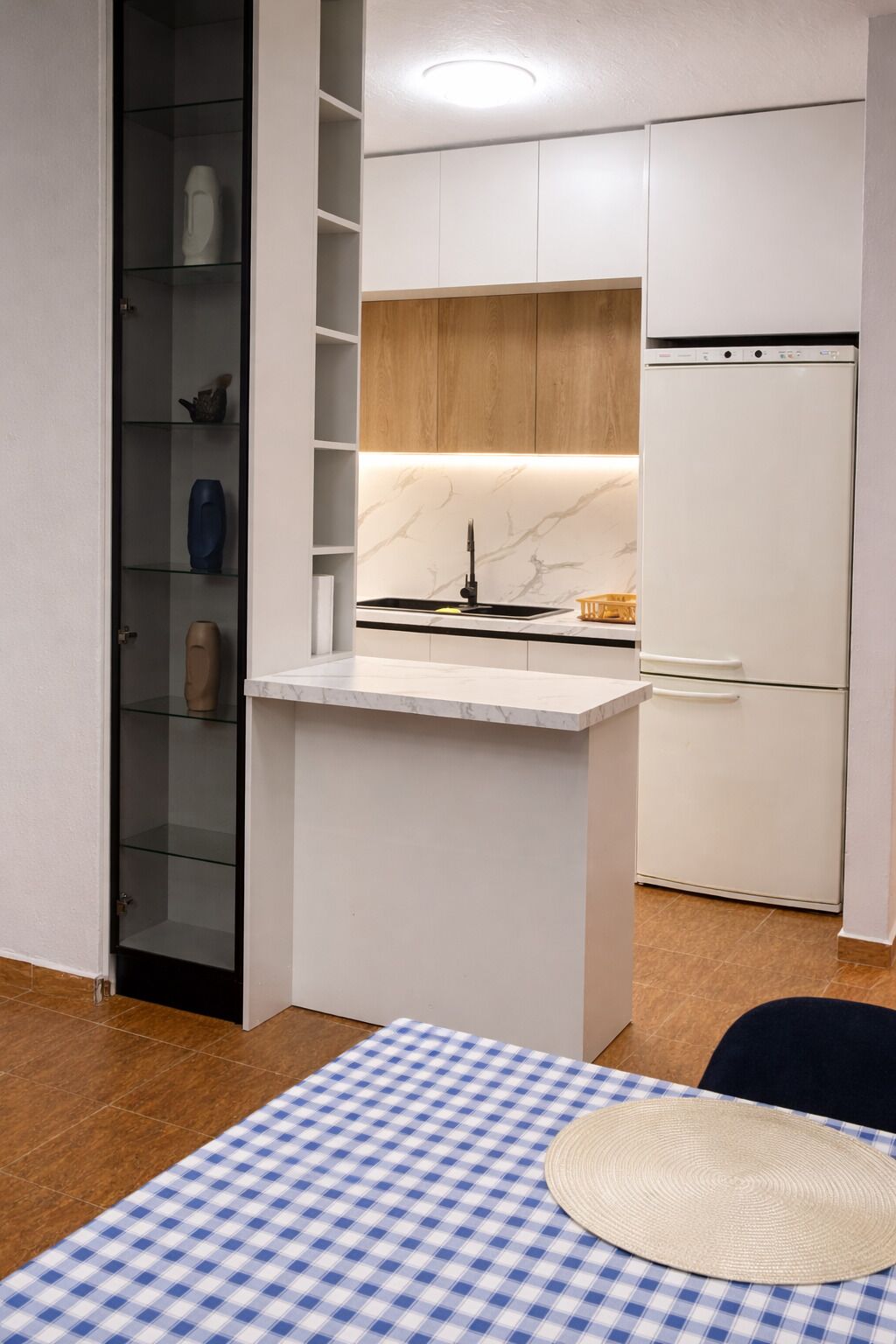 A compact kitchen with a built-in stove, a refrigerator, and a glass-fronted cabinet with decorative items.