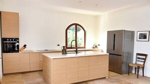 A modern kitchen with a central island, built-in oven, stainless steel refrigerator, and a window with an arched top. - Green Oasis 2 (Lacona)