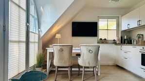A modern kitchen with a dining area, a TV mounted on the wall, and a window with blinds. - Idyllic Thatched Apartment, Schwalbe Wieck Darß, 2 Bedrooms, 2 Balconies, Near Water (Wieck auf Darß)