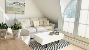 A modern loft bedroom with a sloped ceiling, a white sofa, a white coffee table, and a large window with blinds. - Idyllic Thatched Apartment, Schwalbe Wieck Darß, 2 Bedrooms, 2 Balconies, Near Water (Wieck auf Darß)