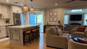 A modern kitchen with a central island, bar stools, a sofa, and a TV in the living area. - Spacious 4 Bed/3 Bath house with pool, fenced backyard, fire pit, covered porch. (St. Simons Island)