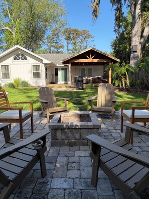 A patio with Adirondack chairs, a stone fire pit, and a covered outdoor seating area. - Spacious 4 Bed/3 Bath house with pool, fenced backyard, fire pit, covered porch. (St. Simons Island)