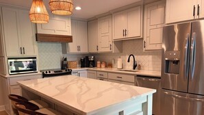 A modern kitchen with white cabinetry, a large island with bar stools, stainless steel appliances, and a herringbone tile backsplash. - Spacious 4 Bed/3 Bath house with pool, fenced backyard, fire pit, covered porch. (St. Simons Island)