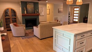 A modern living room with a fireplace, a sofa, a dining area with a white cabinet, and a ceiling fan. - Spacious 4 Bed/3 Bath house with pool, fenced backyard, fire pit, covered porch. (St. Simons Island)
