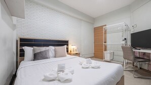 A hotel room with a white brick wall, a bed with white bedding and towels, a desk with a chair, a TV, and a wooden door. - Arun Sa Wad House Lad phrao 4 Guests (Bangkok)
