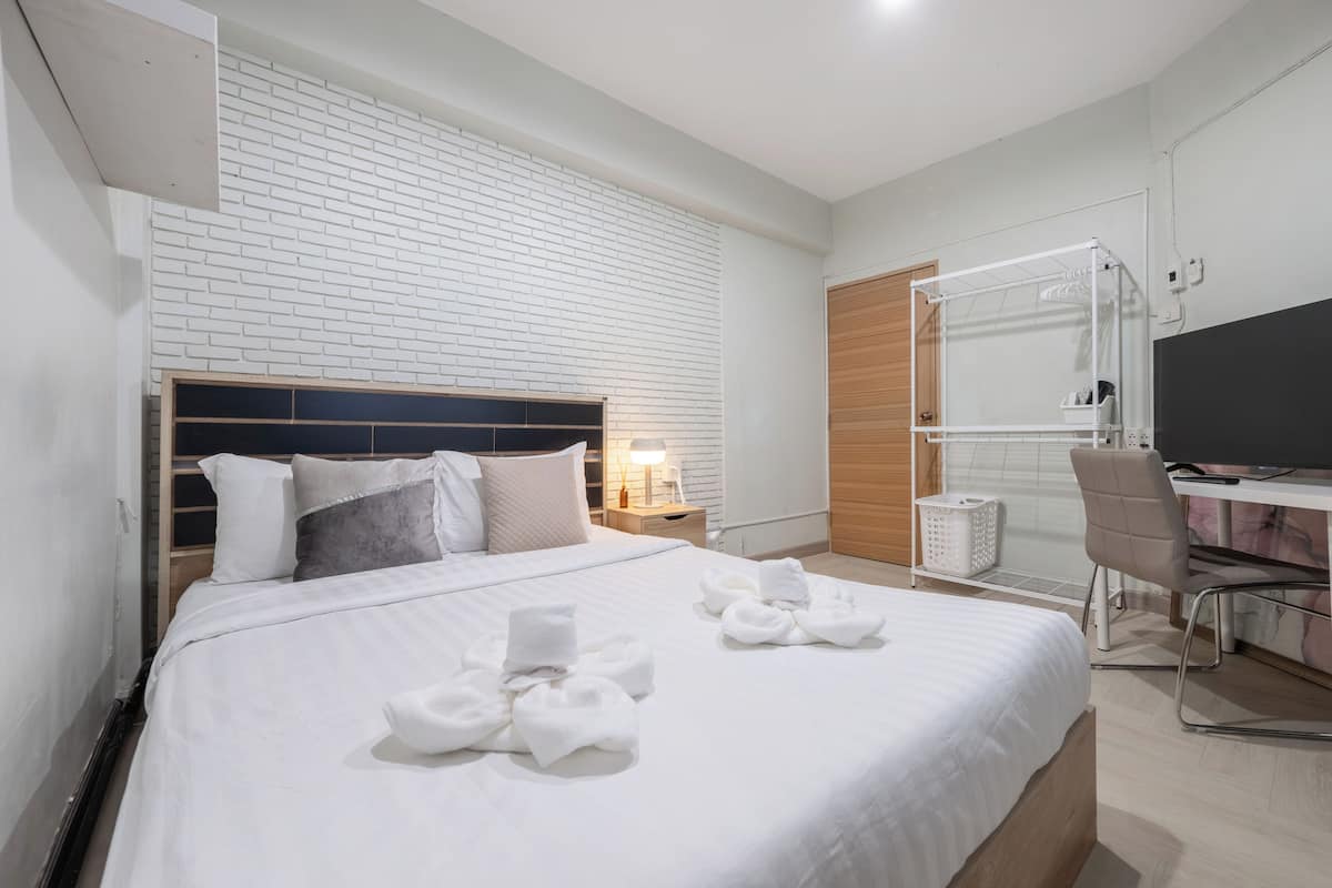 A hotel room with a white brick wall, a bed with white bedding and towels, a desk with a chair, a TV, and a wooden door.