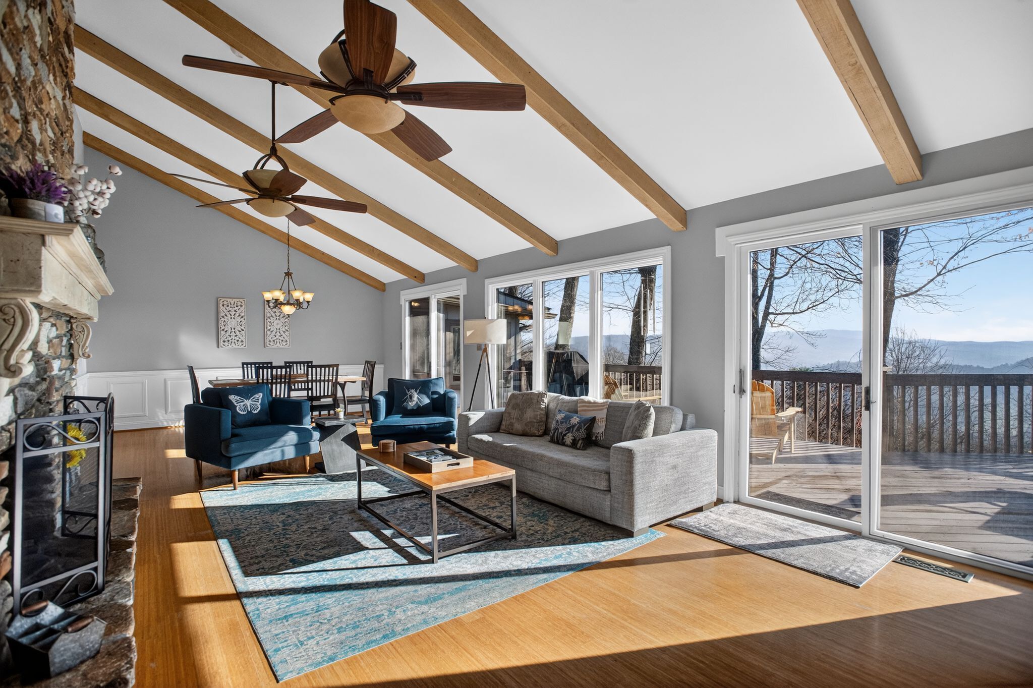 A spacious living room with a fireplace, a ceiling fan, a large window, and a deck with a view of mountains.