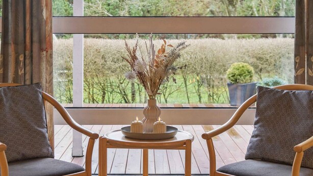 Una habitación con dos sillas de madera, una mesita redonda pequeña y un florero con plantas secas.