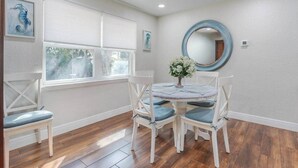 Uma sala de jantar com uma mesa redonda, quatro cadeiras, um vaso com flores, um espelho redondo e uma janela com persianas.