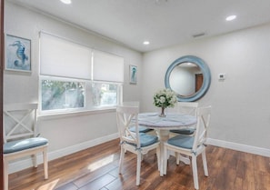 A dining area with a round table, four chairs, a vase with flowers, a round mirror, and a window with blinds. - Coastal Getaway: Near FIT, Downtown & Beach (Melbourne)