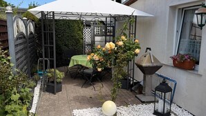 Eine Terrasse mit Tisch und Stühlen, eine Pergola mit weißem Baldachin, ein kleiner Garten mit weißen Kieselsteinen und ein Laternenlicht.