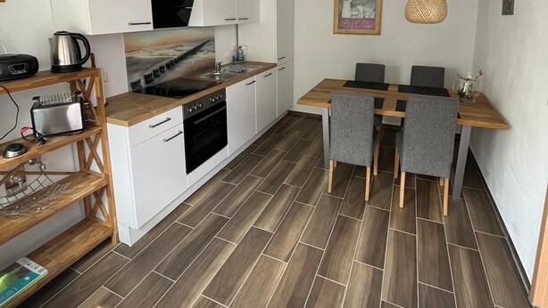 Eine kleine Küche mit weißen Schränken, einem eingebauten Backofen und einem Essbereich mit Holztisch und grauen Stühlen.