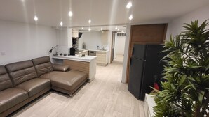 Una sala de estar moderna con un sofá de cuero marrón, una zona de cocina con encimera blanca y un refrigerador negro.