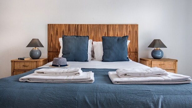 A neatly made bed with blue bedding, two bedside tables with lamps, and a wooden headboard. - Cantinho da Lurdes in Cabanas de Tavira (Tavira)