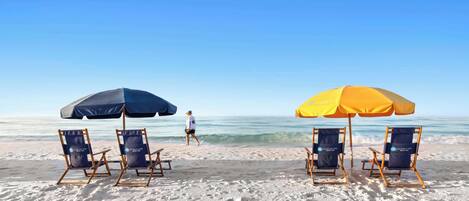 A beach with four lounge chairs, two under blue and two under yellow umbrellas, and a person walking in the distance.