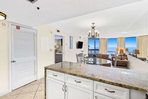A kitchen with a granite countertop, white cabinets, a dining area with a table and chairs, a chandelier, and a view of the ocean. - SunDestin Resort Unit 1517 (Destin)