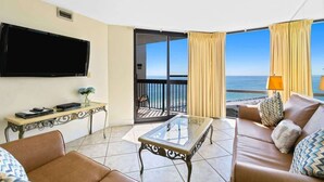 Een hotelkamer met een bank, een glazen salontafel, een flatscreen televisie, een kleine tafel met een vaas, en een balkon met uitzicht op zee.