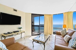 A hotel room with a sofa, a glass coffee table, a flat-screen TV, a small table with a vase, and a balcony with a view of the sea. - SunDestin Resort Unit 1517 (Destin)