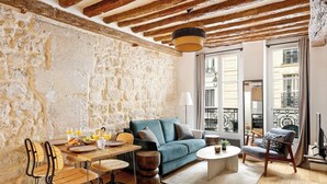 Un salon chaleureux avec un mur en pierre, un canapé bleu, une table à manger en bois avec des chaises, et un balcon doté d’une balustrade en fer forgé.