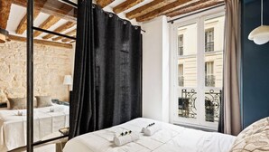 Une chambre avec une grande fenêtre, un lit recouvert de draps blancs, un rideau noir et un mur en pierre.