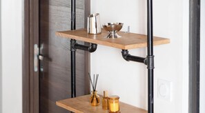 A two-tiered metal pipe shelf unit with a wooden top shelf holding a metal pitcher, a metal bowl, and a small metal container. Below, a lower shelf holds two glass bottles and a reed diffuser. - Loft de charme avec verriere & poutre authentiques (Paris)