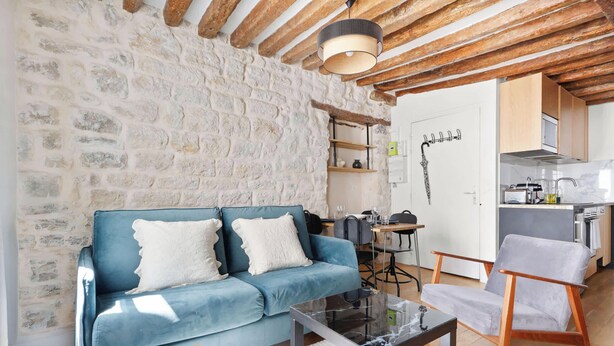 A cozy living area with a stone wall, a blue sofa, a glass coffee table, and a wooden chair. - Loft de charme avec verriere & poutre authentiques (Paris)
