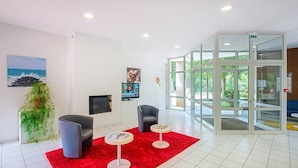 Una hall moderna con tappeto rosso, due poltrone grigie, un tavolino con una rivista, uno schermo televisivo e un ingresso con porta in vetro.