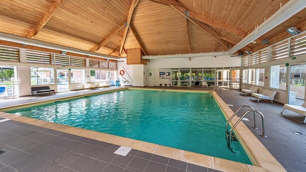 Una piscina interna con soffitto in legno, pavimento piastrellato e finestre illuminate dal sole.