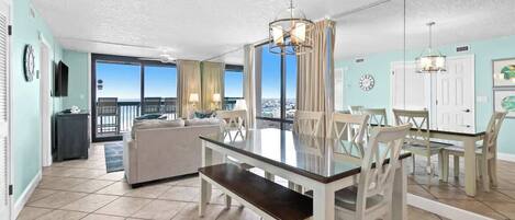 Un vaste espace de vie comprenant une table à manger avec un plateau en verre, des chaises blanches, un canapé et un balcon offrant une vue agréable.
