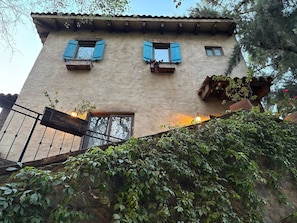 Ein Gebäude mit rustikaler Fassade, mehreren Fenstern mit Fensterläden, einem Balkon mit Pflanzen und einer Treppe zum Eingang.