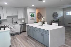 A modern kitchen with white cabinets, a central island, and a dining area with a white table. - Spacious Tampa Retreat  Perfect for Groups + Prime Location (Tampa)