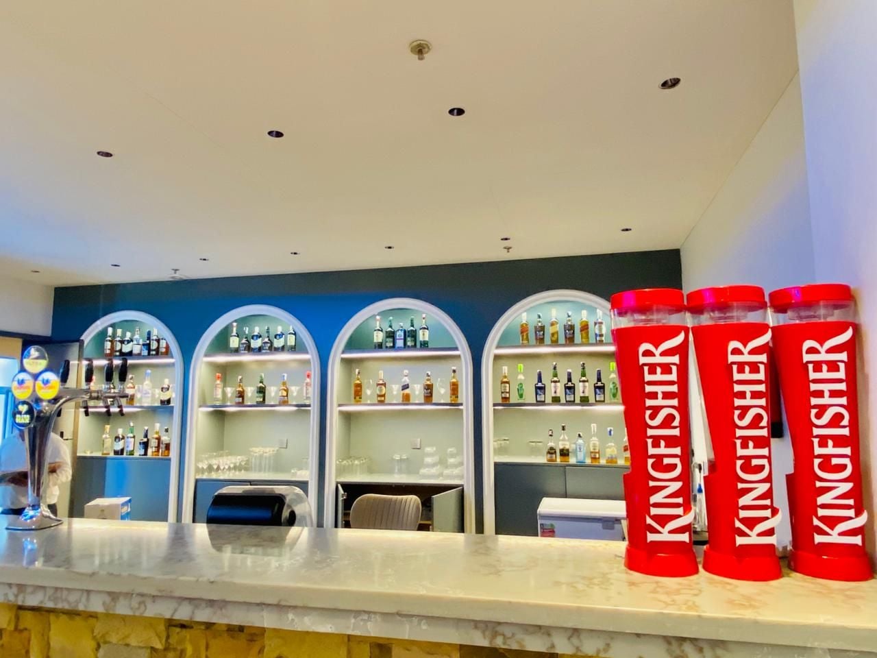 Un espace bar avec des étagères remplies de bouteilles d’alcool, un comptoir et trois bouteilles de la marque Kingfisher, toutes rouges.