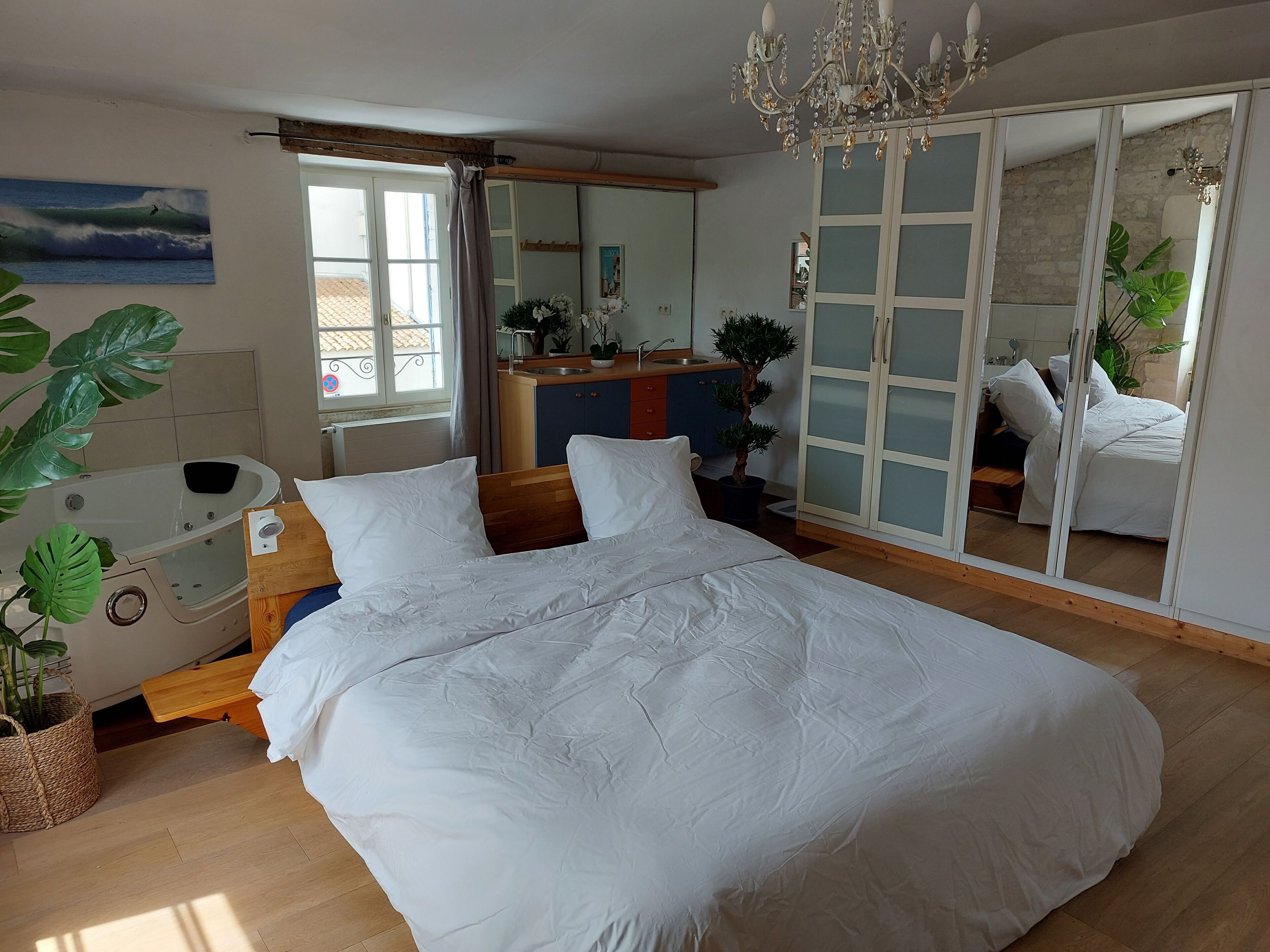 Ein Schlafzimmer mit einem großen Bett, einem Holfnachtkästchen, einem kleinen Tisch mit Pflanze, einer Badewanne, einem Spiegel und Blick ins Freie.