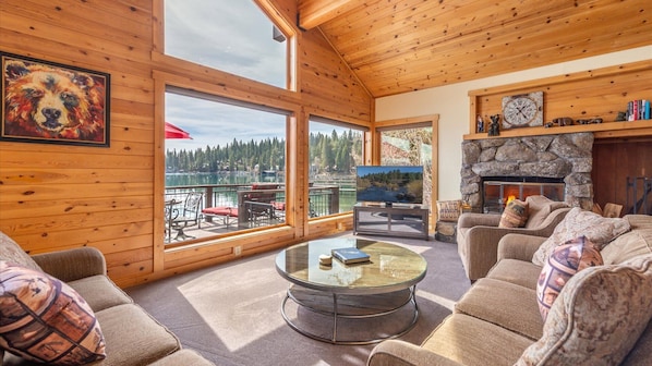 A cozy cabin interior with a stone fireplace, a large TV, and a view of a lake.