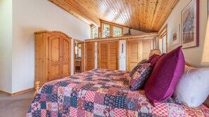 A bedroom with a wooden ceiling, a large bed with a colorful quilt, wooden wardrobes, and a window with a view of trees.